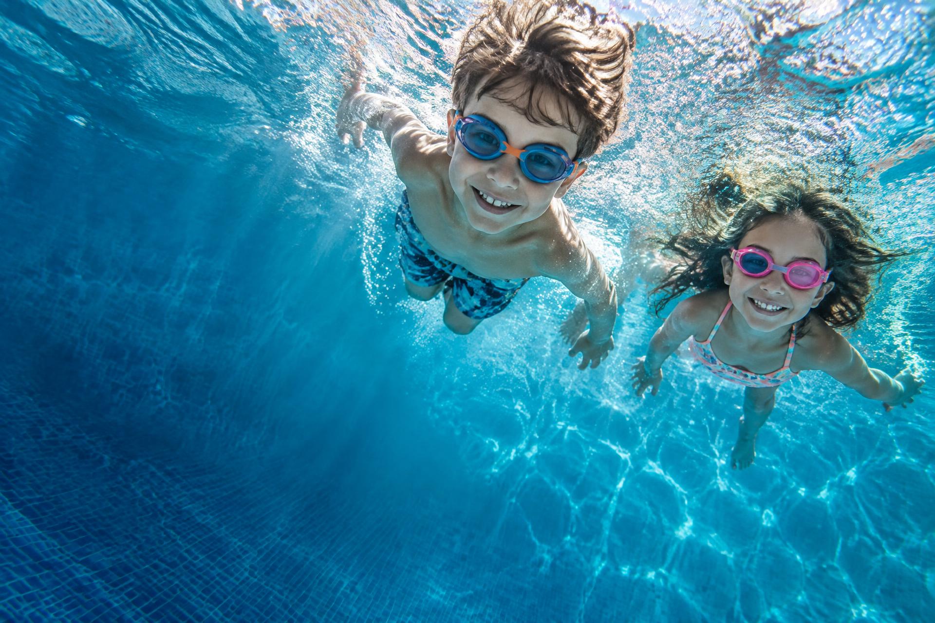 Two children in bright goggles swimming through sunlit aqua pool water